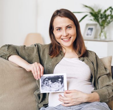 Portrait of happy attractive young woman in homewear sitting on sofa and holding fetal ultrasound image